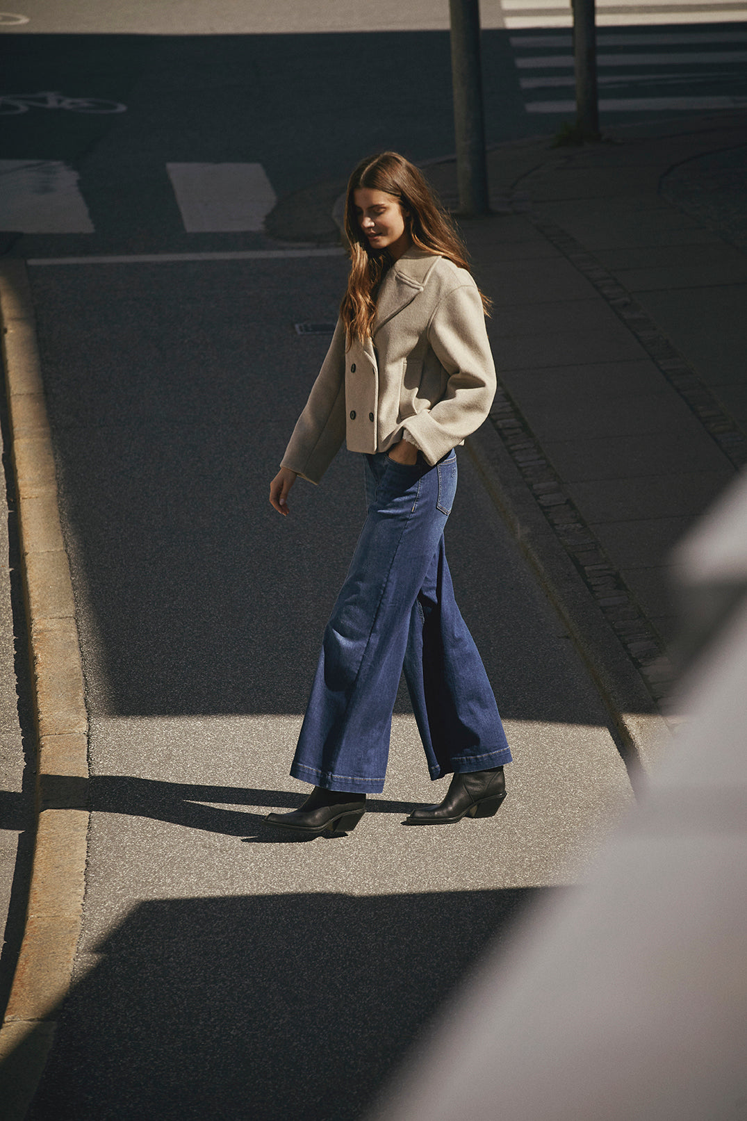 Woman walking on a sidewalk wearing a beige coat and blue jeans.